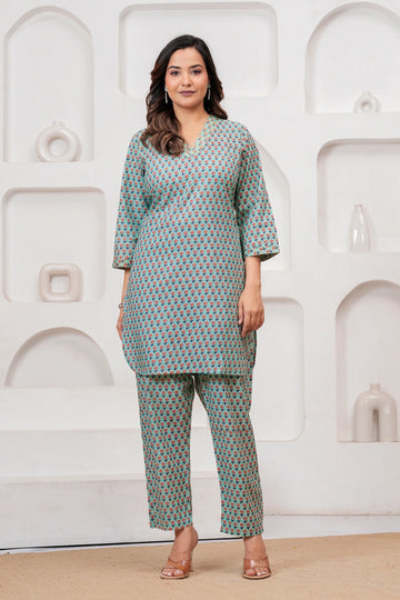A model wearing a teal green co-ord set featuring a tunic and pants with a red and yellow handblock floral pattern against a white decorative backdrop.