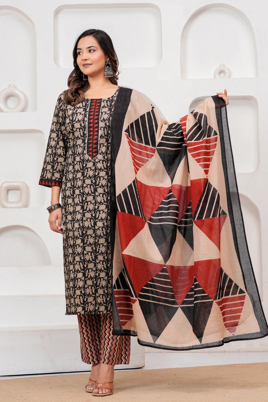 Woman in a black handblock printed cotton kurta with red geometric dupatta and zig-zag printed pants standing against a white textured background.