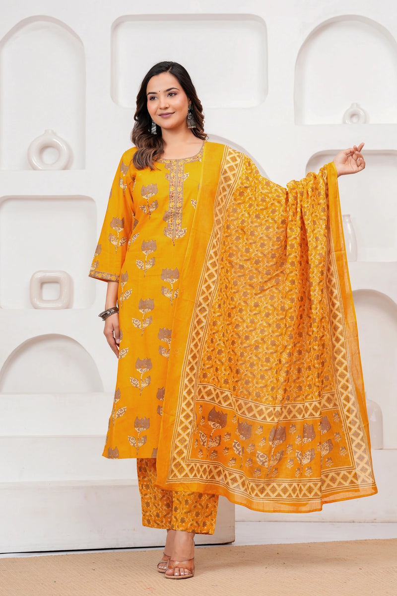 Woman in a mustard yellow floral handblock printed cotton kurta set with a matching dupatta and trousers standing against a minimalist white background.