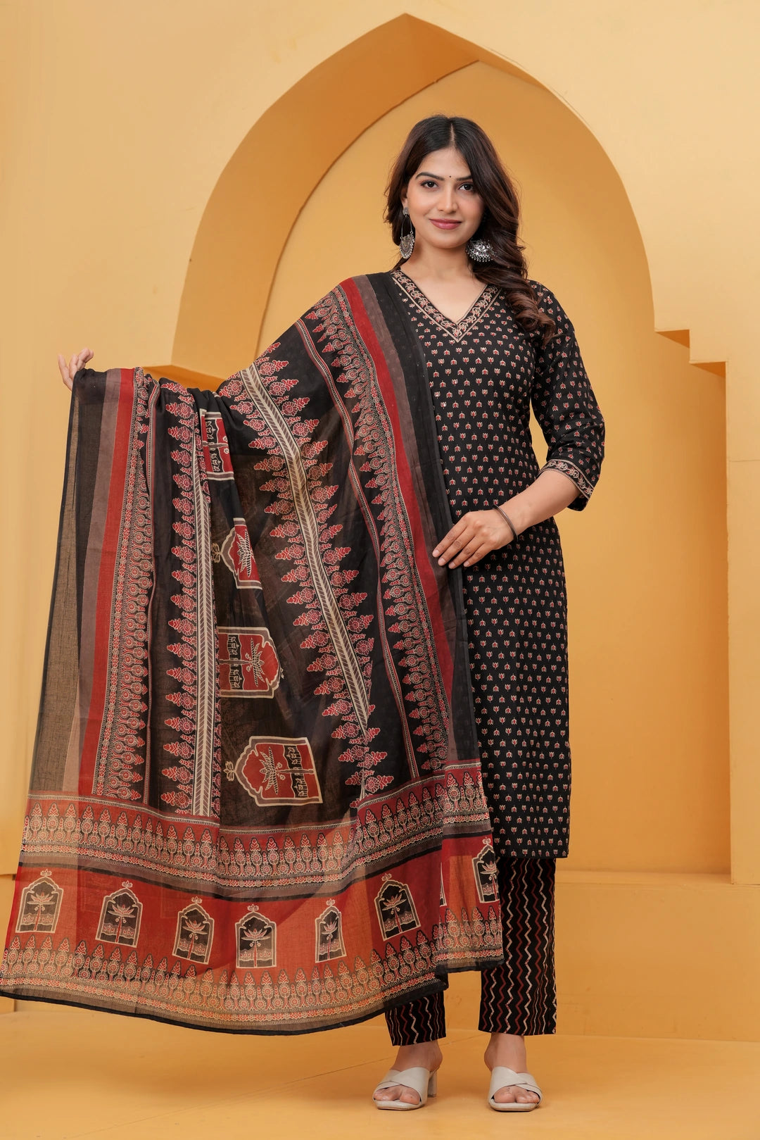 Model posing in a black handblock printed cotton kurta set with a large floral motif dupatta and zigzag print pants against a traditional yellow arch background.