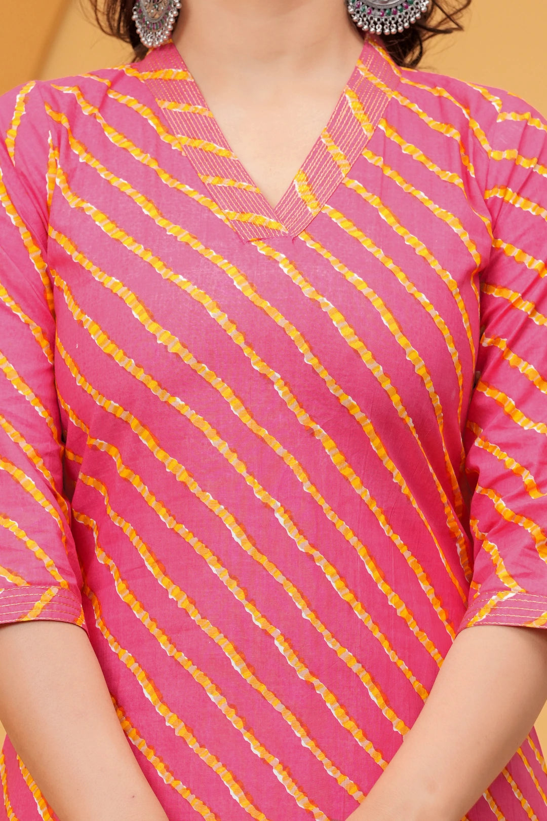 A woman posing in a pink and yellow Leheriya diagonal striped cotton kurta with white straight-cut trousers against a yellow architectural background.