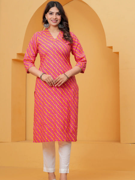 A woman posing in a pink and yellow Leheriya diagonal striped cotton kurta with white straight-cut trousers against a yellow architectural background.